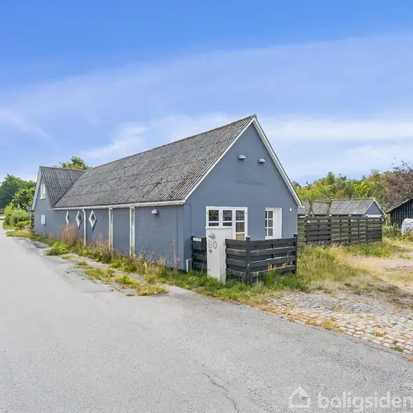 En grå bygning står langs en vej, flankeret af ukrudt og grønt areal. Omkring bygningen er der en indkørsel og en lille hæk. Himlen er blå og klar.