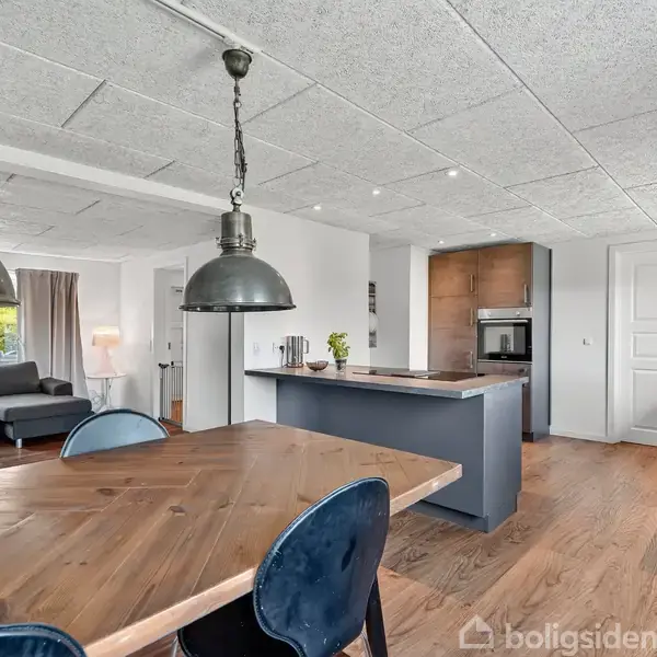 Et moderne køkken med træbord og blå stole, indrammet af to metallysarmaturer. I baggrunden er en stue med grå sofaer, hvidmalede vægge og trægulv.