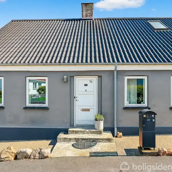 Et gråt hus med hvide vinduer og dør står i en rolig gade. Nummer 16 ses på døren, og en sort postkasse er placeret til højre.