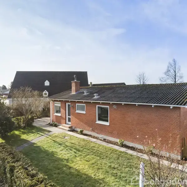 Et rødt murstenshus med fladt tag står i en have. Huset ligger i et boligområde med en græsplæne og buske omkring, under en klar himmel.