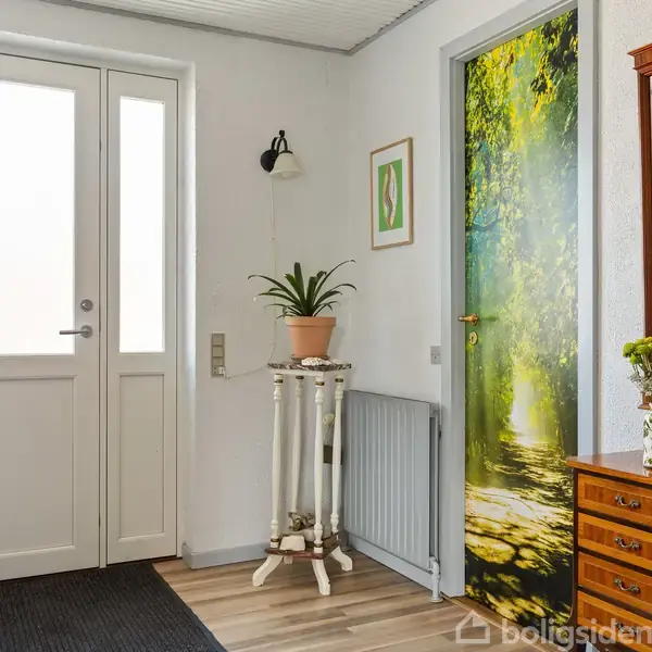 Dør til venstre i en entré, der er lys og indbydende. En plante står på et lille bord, mens et skab med spejl til højre rummer en dekoration. En dør med skovmotiv i baggrunden.