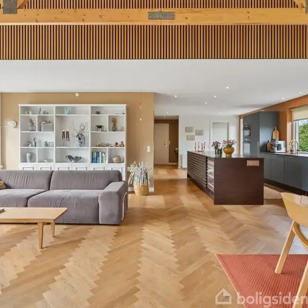 En stue med en grå sofa står foran reoler. Trægulv dækker området, og et åbent køkken med mørke elementer er i baggrunden. Store vinduer giver udsigt til grønne omgivelser.