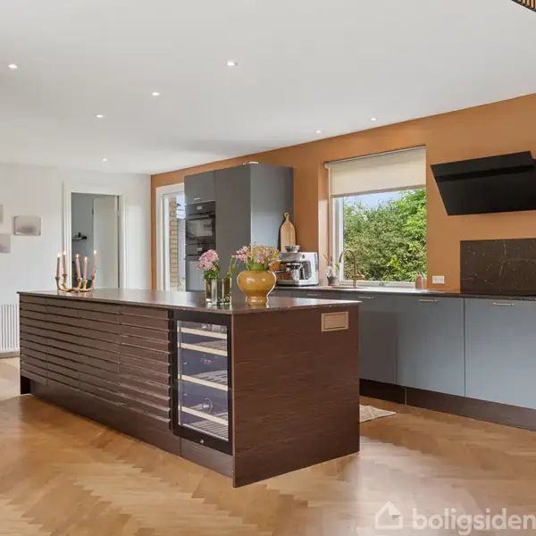 Køkken med mørk træø indeholder indbygget vinkøler og dekoreret vase. Skabe langs væggen under vinduet overfor viser køkkenudstyr. Rummet har trægulv og orange væg.