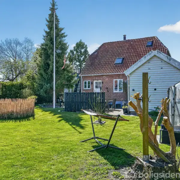 Et rødt teglhus står i en have med græsplæne. Til højre ses et drivhus, mens en hængekøje hænger i midten. Baggrunden består af træer og blå himmel.