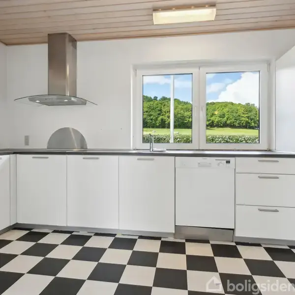 Et moderne køkken med hvide skabe står tomt. Køkkenet har et sort-hvidt skakternet gulv og udsigt til grønne træer gennem et stort vindue.