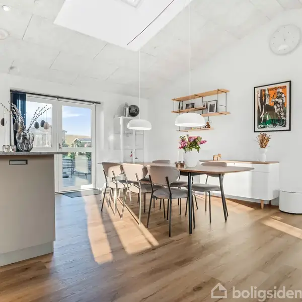Et lyst og moderne køkken-alrum med træbord og stole, blomster, vægkunst og belysning. Store vinduer slipper naturligt lys ind, og der er adgang til en udendørs terrasse gennem glasskydedøre.