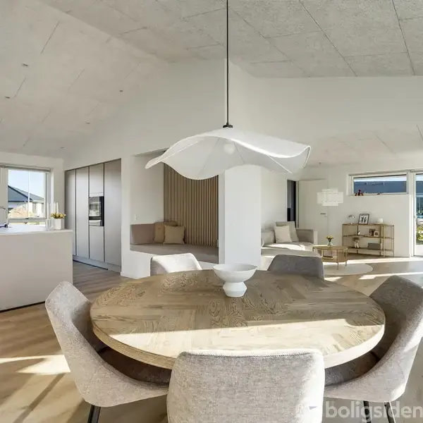 Et rundt spisebord med stole står centralt i et lyst og moderne køkken-alrum. Store vinduer giver udsigt til haven, og en smuk lampe hænger over bordet.