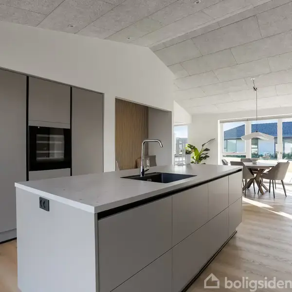 Køkken med køkkenø, indbygget ovn og moderne skabe står i et lyst rum. Spisebord og stole ses ved et stort vindue, der giver udsigt til en terrasse med blå tag.