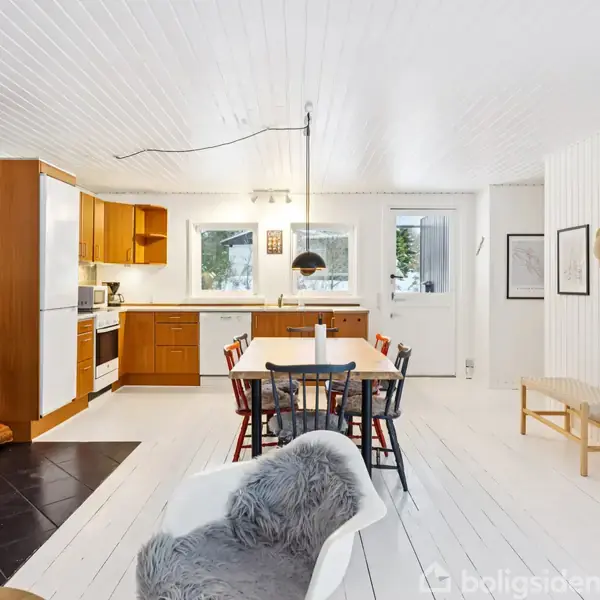 Brændeovn opvarmer stue-køkken-alrum med hvidmalede trævægge og lofter. Langt spisebord fyldt med stole, lys fra vinduer. Køkken med træskabe i baggrunden. Oval vægdekoration.