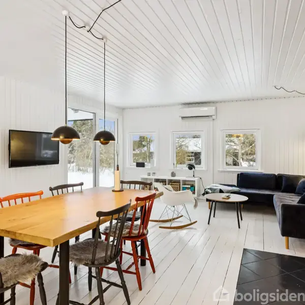 Et lyst stueområde med spisebord og stole i front. Tre pendellamper hænger over bordet. I baggrunden står en mørk sofa ved siden af vinduer. Brændeovn i højre hjørne.