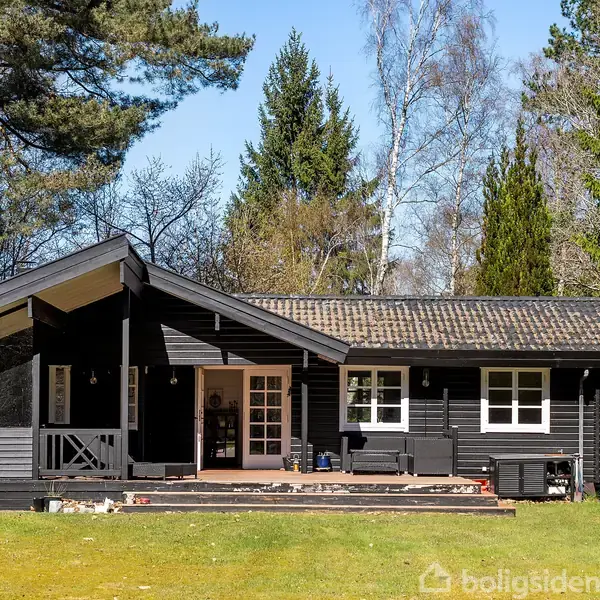 Et sort træhus med terrasse står i en lysning omgivet af høje træer. Huset har store vinduer og en overdækket veranda. Der er græs og skov som omgiver huset.