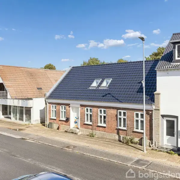 Rækkehuse med forskelligt farvede tage står langs en vej. En bil holder parkeret ved gaden, og der ses blå himmel med få skyer i baggrunden.
