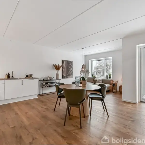 Et rundt spisebord står med stole i en rummelig stue-køkken-alrum. Lyse træmøbler, hvidt køkken og en åben terrassedør skaber en lys og indbydende atmosfære med udsigt til sneklædt have.