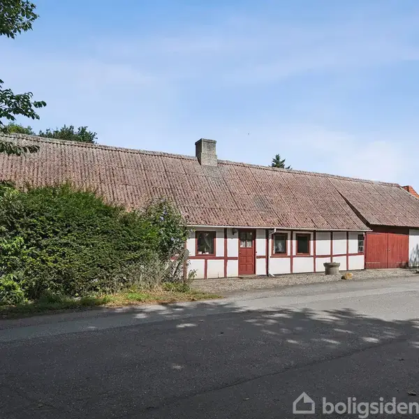 Gammelt bindingsværkshus med røde og hvide felter, set fra en stille vej. Taget er af falmet teglsten. Omgivet af træer og buske i et roligt landligt område.