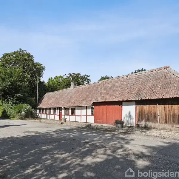Længere bindingsværkshus med stråtag og røde døre står i et solrigt, landligt område ved en vej. Omkring huset er der grønne træer og buske.