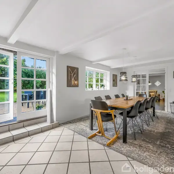 Lang spisebord med sorte stole står på et tæppe i en lys spisestue. Store vinduer og dobbeltdøre i glas giver udsigt til grøn have.