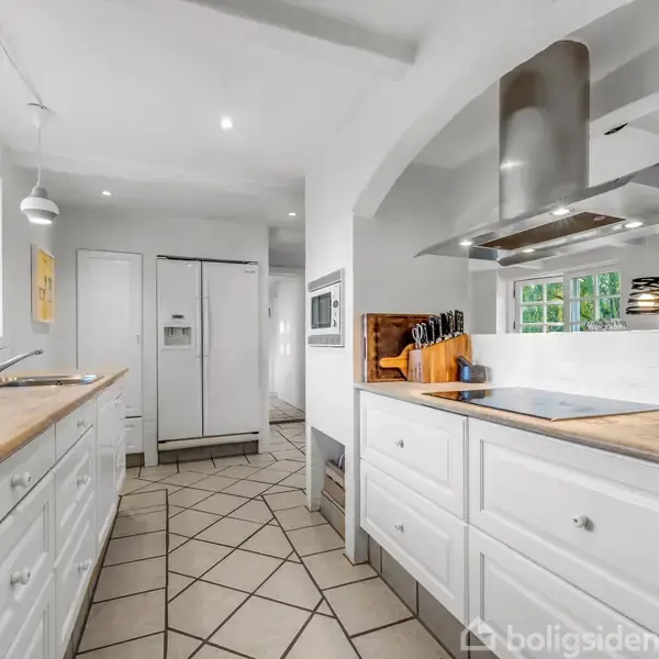 Et moderne køkken med hvide skabe, hvor knivblok og emhætte flankerer kogepladen. Køkkenet har lyst trægulv og fliser samt vinduer, der giver naturligt lysindfald. Ingen tekst fundet.