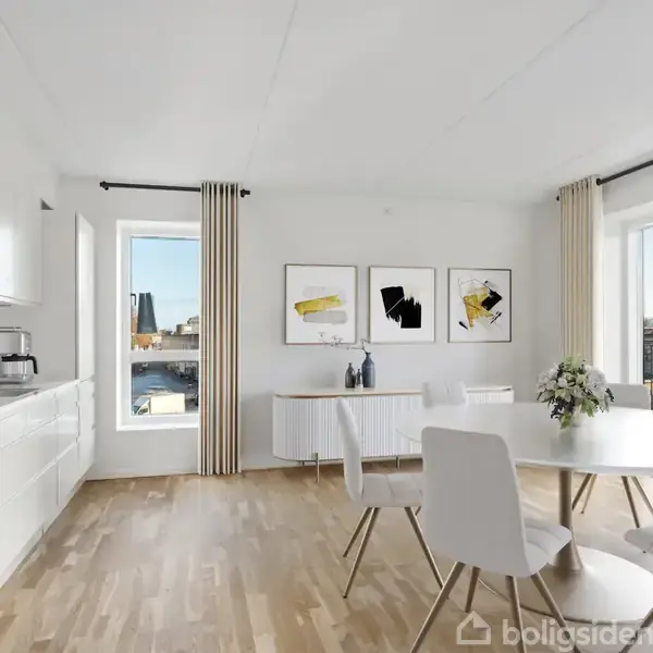 Et lyst køkken-alrum med hvidt køkken, rund spisebord med fire stole, trægulv og moderne kunst på væggen. Store vinduer med udsigt og lyse gardiner skaber en åben atmosfære.