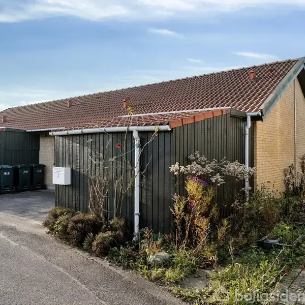 En villa med rødt tegltag står ved en vejkant, omgivet af en have med buske og blomster. Til venstre ses affaldscontainere op ad en grøn væg.