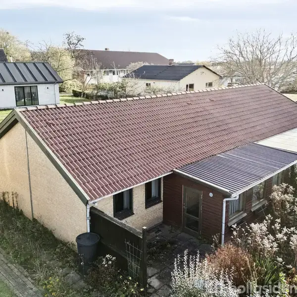 Huset har et rødt tegltag og lysegule mure, omgivet af have med buske og planter. I baggrunden er der flere huse og træer.
