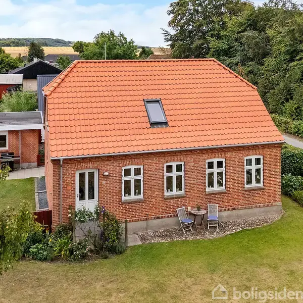 Rødt murstenshus med skråt, rødt tegltag og ovenlysvindue. Foran huset står et cafébord med to stole på en lille terrasse. Omgivet af græsplæne og træer i landlig område.