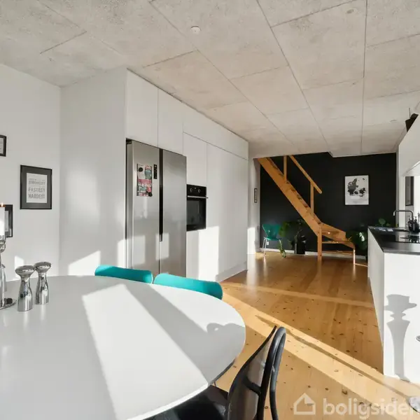 Rundt spisebord med lysestager og stole står i en moderne, lys køkken-stue med trætrappe til førstesal. Væggene har sort-hvide billeder og der er et åbent køkken i baggrunden.