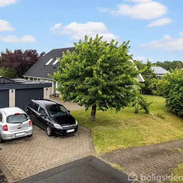 Et parcelhus med tilhørende garage og indkørsel ses omgivet af grønne træer og en græsplæne. To biler holder parkeret på den stenbelagte indkørsel. En til salg-skilt står ved siden af postkasser.