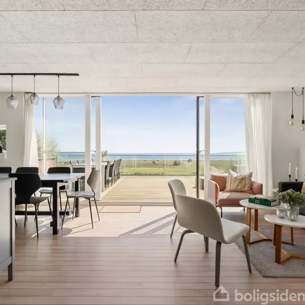 Stue med moderne møbler, åben til køkken, udsigt til havet gennem store glasdøre, trægulve og minimalistisk indretning.
