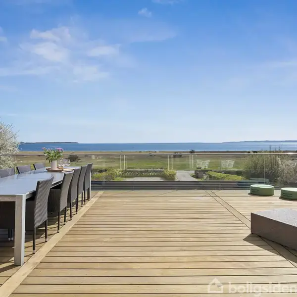 Et langt bord med sorte stole står på en træterrasse. Bordet er pyntet med planter. Terrasseområdet har udsigt over en græsmark og havet i horisonten under en blå himmel.