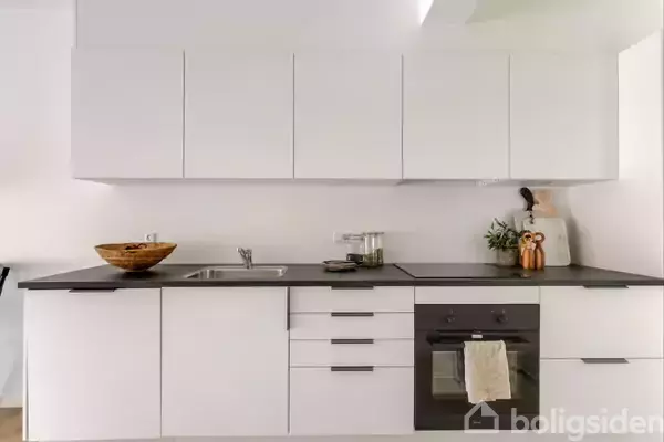 Et moderne køkken har hvide skabe, en sort bordplade med en vask, kogeplader og en indbygget ovn. En skål, planter og køkkenredskaber er placeret på bordet.