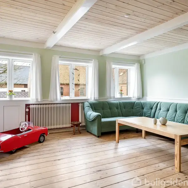 En stue med en grøn hjørnesofa står ved et træbord. Rummet har trægulve, tre vinduer med hvidt lysindfald, et rødt legetøjsbil placeret foran en radiator og en lampe på en kommode.