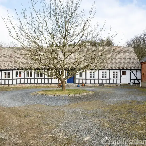 Bindingsværkshus står i centrum omkranset af grusvej, omgivet af træløse grene. Rødt murstensbygning ses til højre. Himlen er skyet. Ingen tekst er til stede.