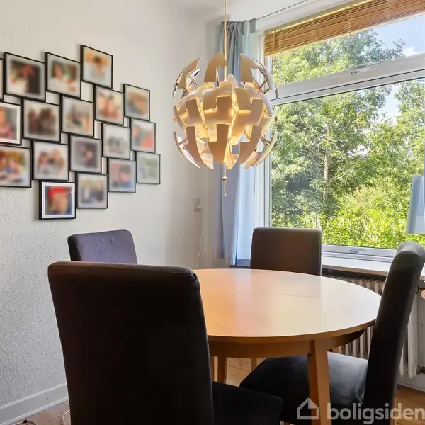 Rundt spisebord med fire stole står i en lys spisestue. En moderne lampe hænger over bordet, og en væg har en collage af slørede billeder. Stort vindue med grønt udsyn.