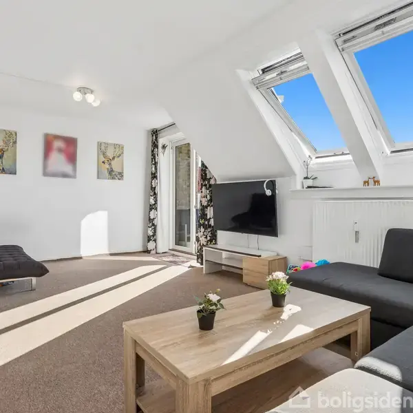 Stue med gråt hjørnesofa står på brunt tæppe. Et træsofabord med planter. Fladskærm foran skråvinduer. Malerier hænger på hvide vægge. Døren fører til balkon.