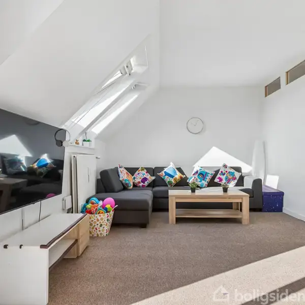 En lys stue med grå hjørnesofa fyldt med farverige puder. Foran sofaen står et træbord. Et stort TV hænger på væggen. Legetøjskurve og loftsvinduer skaber en hyggelig atmosfære.
