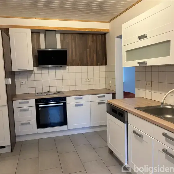 Køkken med hvide skabe og indbygget ovn under en sort emhætte. En sort opvaskemaskine ved siden af en stålvask. Flisebeklædning på væggene i rummet.