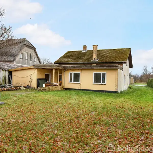 En gulmalet, en-etagers hus står på en stor græsplæne med få spredte træer. Huset har skråt tag og skorstene, omgivet af en rolig landskabelig have.