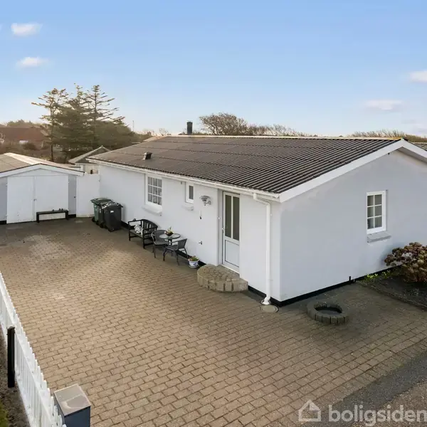 En hvid, én-etagers hus står stille på en brolagt indkørsel, omgivet af en lav hæk og græsplæne foran en garagebygning under en blå himmel.
