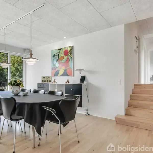 Et spisebord med sorte stole står i en lys spisestue ved siden af et stort vindue med udsigt til natur. En farverig plakat hænger over en skænk, og en trappe leder opad.