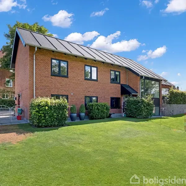 En rødstensbygning med sort tag står stille i en grøn have, omgivet af buske, med en lyseblå himmel og få skyer i baggrunden.