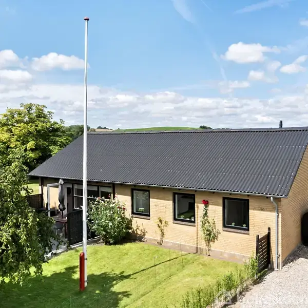 En gul murstensvilla med sort tag står stille i en velplejet have med frodig græsplæne og buske. En høj flagstang og grusindkørsel er synlige i forgrunden.