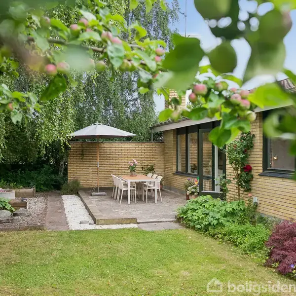 Terrassebord med stole står under en parasol ved siden af et gult murstenshus. Haven er frodig med græsplæne, blomster, og træer, der skaber en hyggelig udendørs atmosfære.