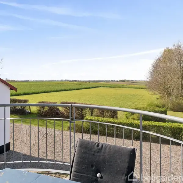 Balkon med metalgelænder omgiver et bord og stol, set mod en udsigt over åbne, grønne marker og træer under en blå himmel. En bygning vises til venstre.
