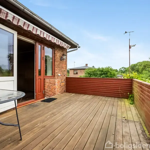 Bord står på en træterrasse ved et rødstenshus med åbne døre. Terrassen er omgivet af lave træhegn, og der er udsigt til grøn vegetation og huse i baggrunden.