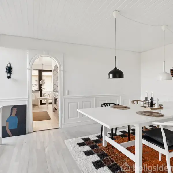 Spisebord med fem stole står i en lys spisestue med kunst på væggene. En stor gulvlampe og maleri står ved en lysbue, som fører til et andet rum.