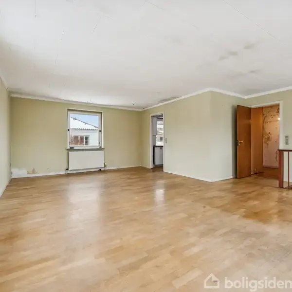 En lys stue med trægulv og beige vægge. Et åbent område med store vinduer til venstre, en radiator og døråbning til et køkkenområde til højre.