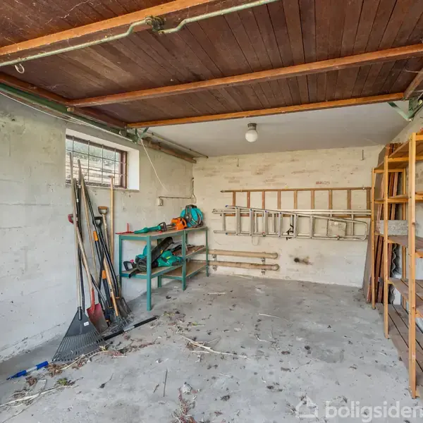 Garage indrettet som værksted med arbejdsbænk, værktøj på væggen og hylder på siden. En vask er placeret i hjørnet. Betongulv og træloft præger rummet.