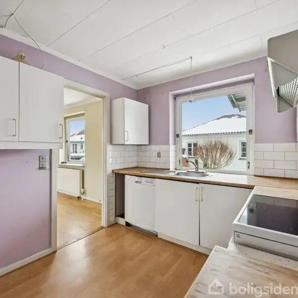 Et lille køkken med hvide skabe og træbordplader. Et vindue over vasken giver udsigt til hustage. Rummet har lilla vægge og lyst trægulv, tæt forbundet til en gang.