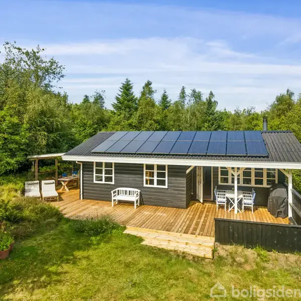 Et sort træhus med solpaneler på taget står midt i en grøn have og skovområde. Der er en terrasse med hvide havemøbler foran huset.