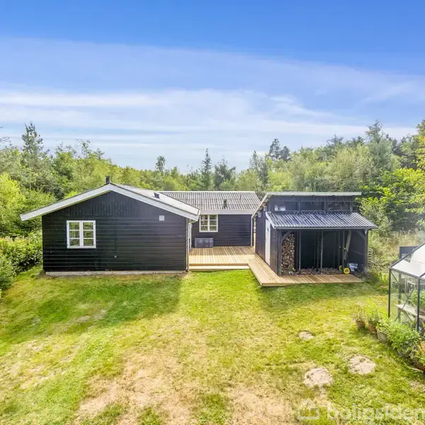 Sort træhus står stille blandt grønne træer og græsplæne. Et drivhus ses i højre kant af billedet. Himmel er klar med få skyer.
